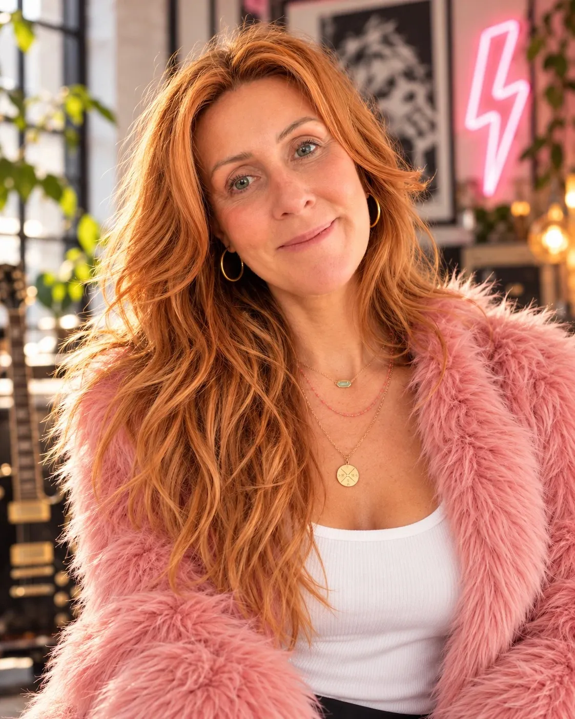 Kenda Hansen in a pink faux fur coat and white tank top, long copper hair, gold hoops and layered necklaces, a pink neon lightning bolt behind her