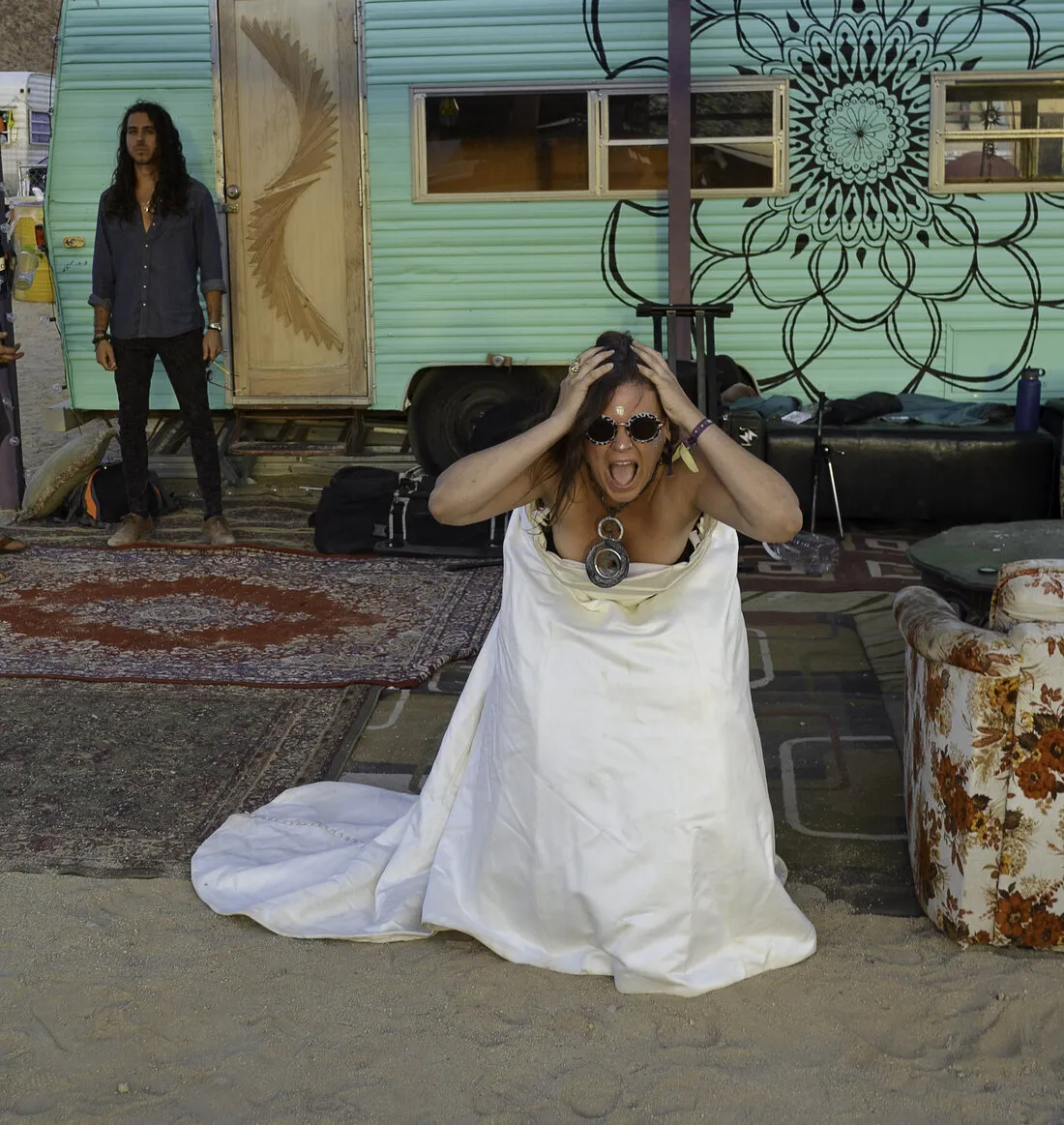 A woman in The Divorce Dress kneeling on a rug in the desert, hands on her head, mid scream
