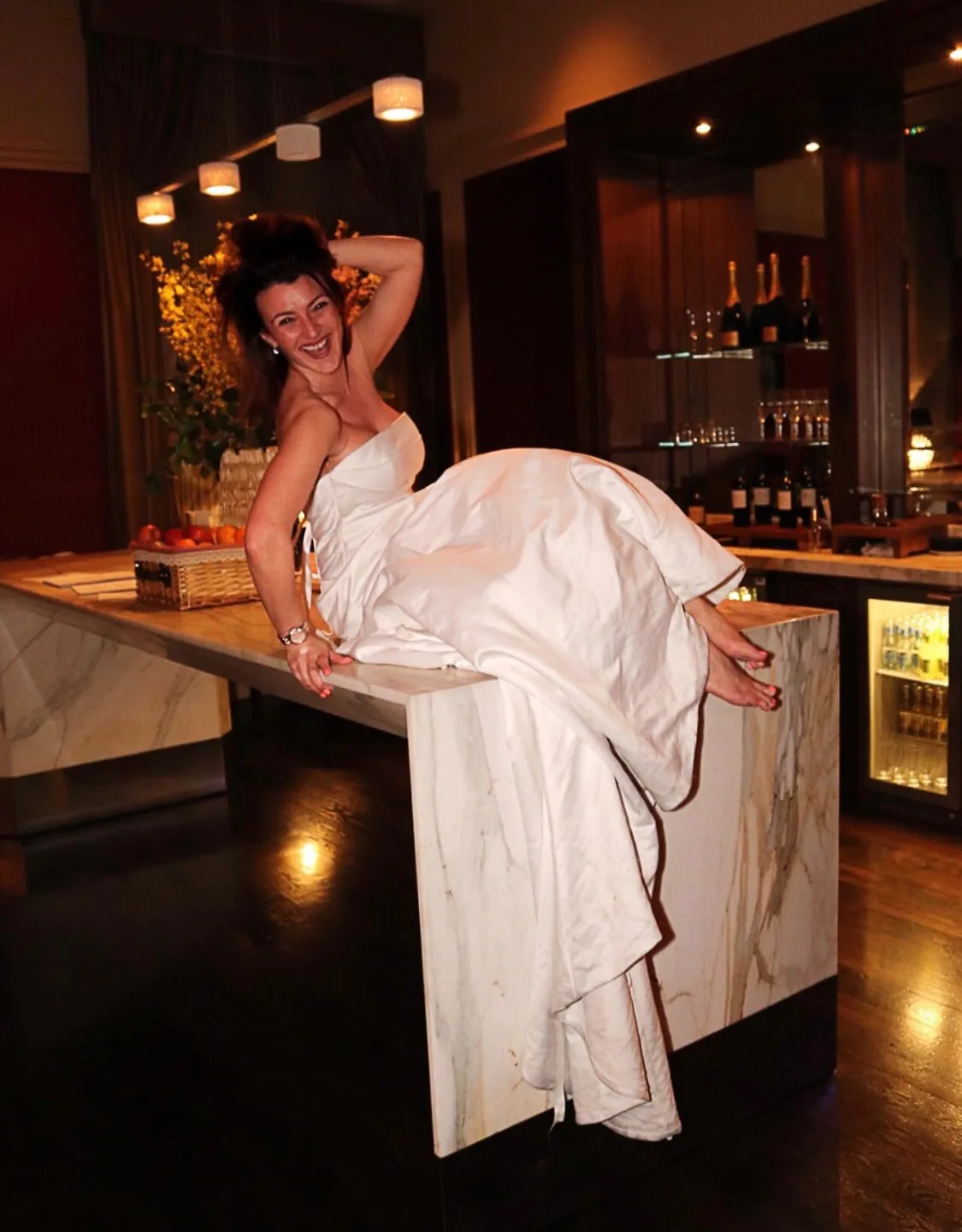 Kenda laughing in The Divorce Dress, perched barefoot on a marble bar at night