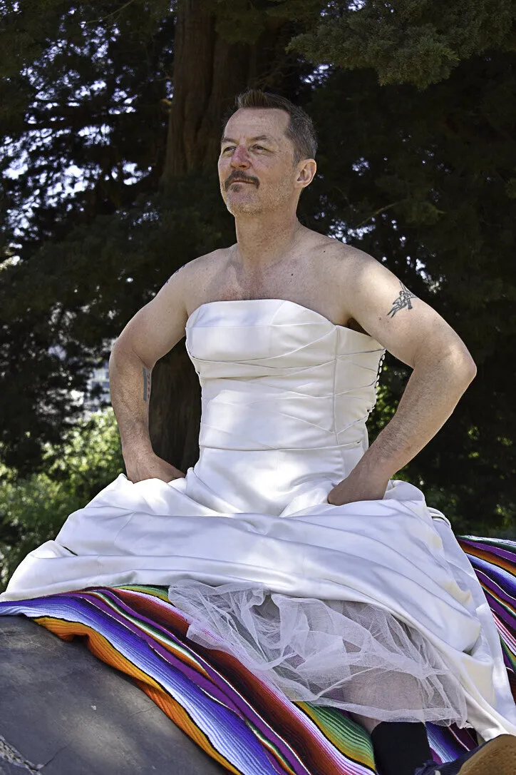 A man with a mustache wearing The Divorce Dress, sitting on a striped blanket under the trees
